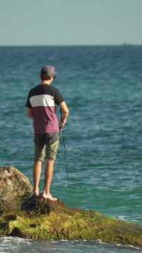 Man fishing ocean, standing on rocks with a rod in the clear blue sea on a sunny day. Vertical video.