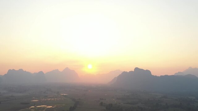 Aerial view of dramatic limestone karst mountains at sunrise in Vang Vieng, Laos. Golden morning light illuminates the hazy rural valley and rice fields