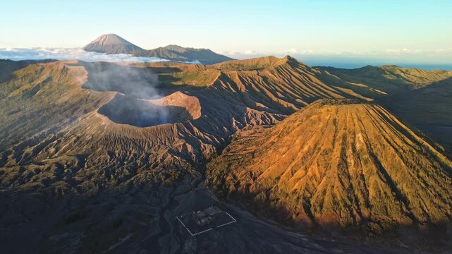 Drone slowly pulls away from the Mount Bromo volcanic landscape in vibrant golden-yellow morning light, revealing the volcanic peaks and clear blue sky, Indonesia