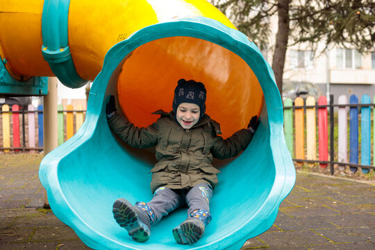 Cheerful little boy enjoys playtime at a colorful fenced playground, laughing on a round nest swing and sliding down a bright tube slide outdoors