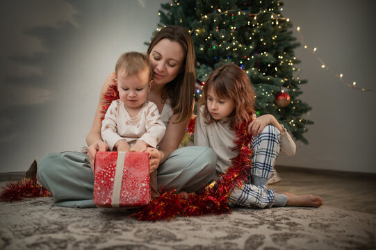 Mother and daughters opening presents under the tree, New Year and Christmas celebrations. Gift wrapping real people, real emotions