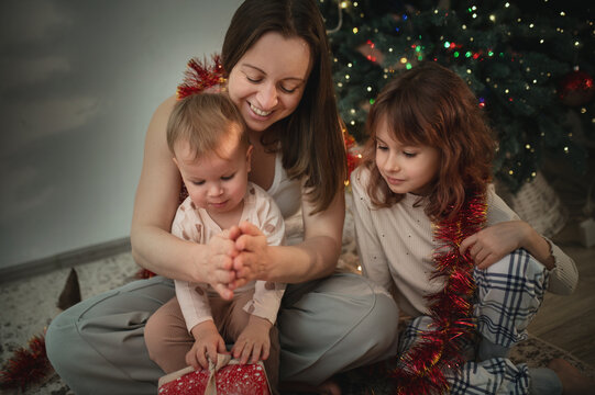 Mother and daughters opening presents under the tree, New Year and Christmas celebrations. Gift wrapping real people, real emotions