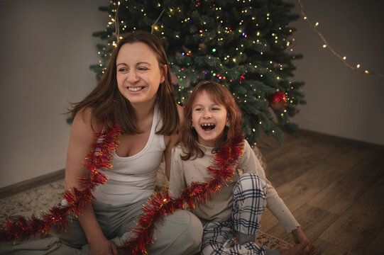 A mother and daughter celebrate the New Year under a Christmas tree with garlands and lights. A happy, emotional photo of real people, a homey atmosphere.