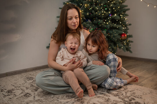 family of 2 children, mom are celebrating New Year's and Christmas under Christmas tree at home. Children crying under the Christmas tree, difficult parenting, real life, childish hysteria