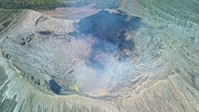 Close drone flight over Mount Bromo crater revealing the smoking crater floor, rugged interior and tourist ascending the stairs toward the crater rim, Indonesia