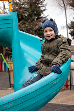 Cheerful little boy enjoys playtime at a colorful fenced playground, laughing on a round nest swing and sliding down a bright tube slide outdoors