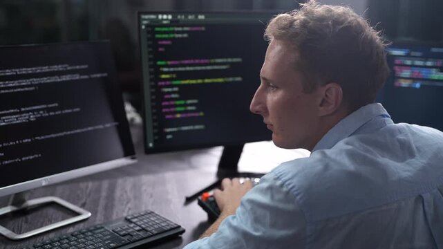 Male software developer coding on multiple monitors in dark office. IT programmer analyzing global network data on screen. Corporate cybersecurity technology and software development industry concept