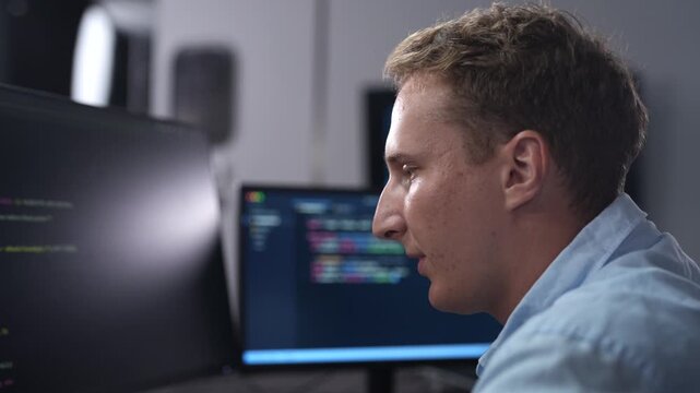 Male software developer coding on multiple monitors in dark office. IT programmer analyzing global network data on screen. Corporate cybersecurity technology and software development industry concept