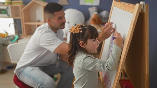 Male teacher crouching beside young girl drawing on easel with marker, pointing at whiteboard and smiling inside daycare building; calm focus.