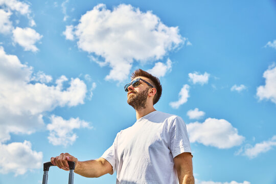 Young Man with Suitcase Standing Outdoors Against Blue Cloudy Sky