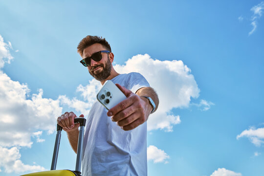 Young man holding smartphone and suitcase against blue sky