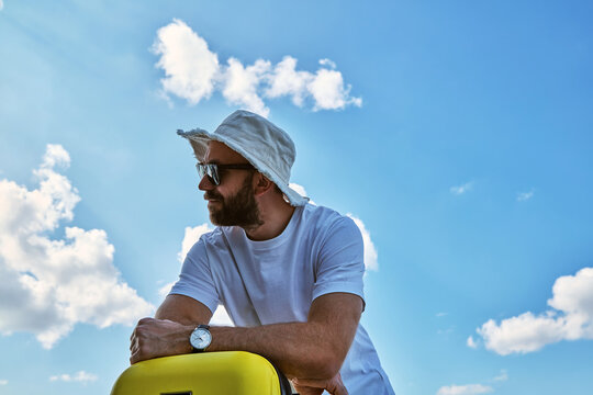 Young man with yellow suitcase against blue sky waiting for departure during summer vacation