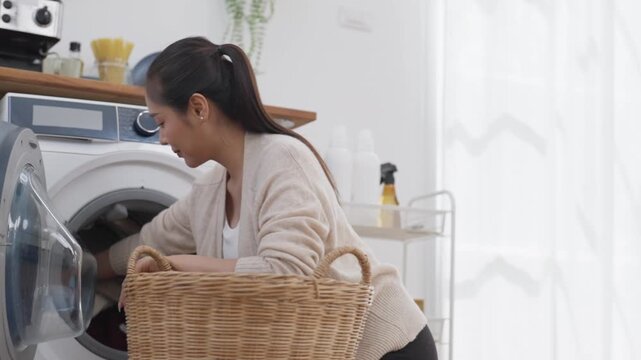 Young adult Asian woman doing laundry with wicker basket beside front load washing machine in clean bright home kitchen appliance interior calm domestic chore scene