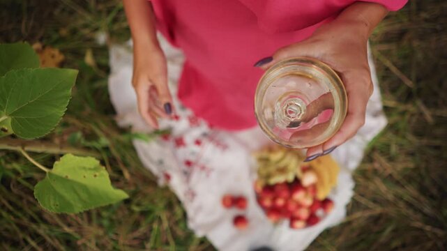 Close up of woman in pink holding empty glass jar with dark blue nails over grass and fruit. scene includes strawberries apples bananas on white cloth outdoors under natural light
