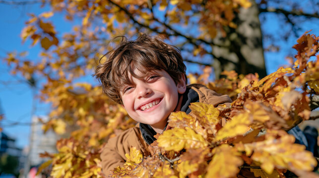 Close up of a smiling young boy hanging playfully from a low tree branch in an autumn park, face framed by golden yellow leaves, tousled hair and warm casual clothing, joyful expression, shallow dep