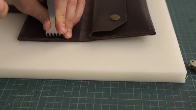 Leatherworker punching stitching holes into a handmade brown leather wallet using a prong chisel and a mallet in a workshop