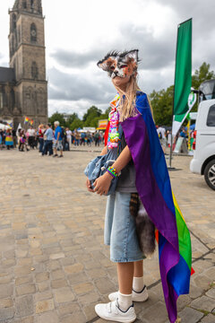 Masked therian girl in colorful rainbow cape poses during CSD celebration in Magdeburg, Germany, with cathedral behind. Scene reflects youth subculture, fandom, identity, pride, creativity, community