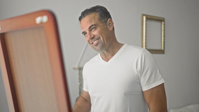 Man smiles into a standing mirror while holding a makeup brush and wearing a white v neck t shirt, casual grooming pose in studio; confidence wellness.