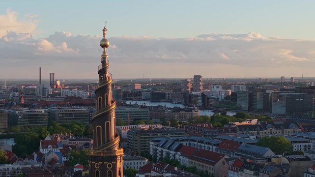 Aerial footage circling the historic belfry of Our Saviour's Church Belfry (Vor Frelsers Kirke) as warm evening sunlight illuminates Copenhagen's rooftops, architecture, and urban skyline.