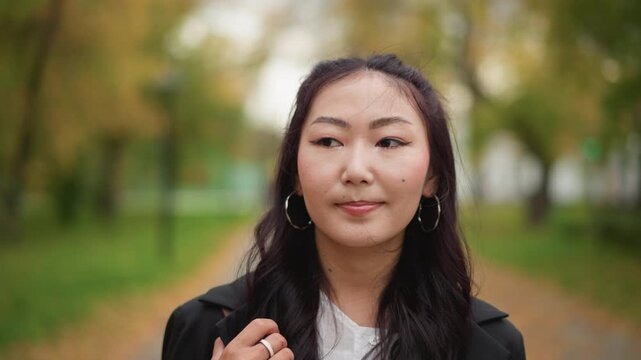 Close up of young woman with long dark hair wearing large hoop earrings and holding lapel of her black coat in outdoor park setting. scene features soft natural light green grassy areas