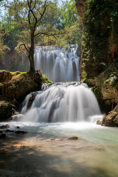 Vista vertical en plano medio de una cascada natural de dos niveles cayendo violentamente sobre rocas en un r&iacute;o rodeado de &aacute;rboles y vegetaci&oacute;n frondosa en un d&iacute;a soleado. Monasterio de Piedra.