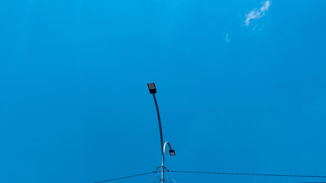 Street light pole standing tall against a bright blue sky background with minimal clouds and electrical wires below providing urban infrastructure lighting