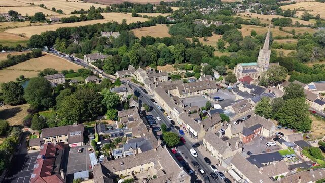 Aerial drone orbit around St John the Baptist Church in Burford, showcasing the Church of England spire, historic stone roofs, and Fulbrook Road stretching toward the Oxfordshire countryside