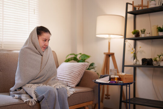 Sick Asian woman wrapped in blanket shivering with cold fever on couch at home