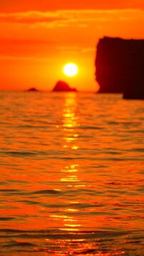 Fiery orange sunset illuminates rolling tide near black silhouettes of coastal rock formations in serene tropical water. Vertical fixed low angle shot.