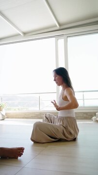 Young female yoga instructor leading a private session and teaching breathing techniques in a bright studio with panoramic windows, promoting mindfulness, relaxation and holistic wellness.