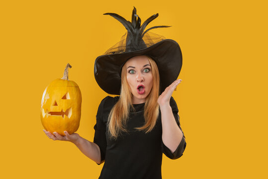 Woman wearing a black witch costume and a pointed hat, holding a glowing carved Halloween pumpkin and showing a shocked or surprised facial expression on a yellow background