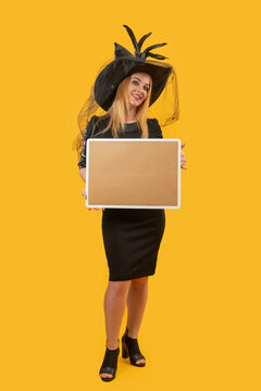 Young smiling woman dressed in a black witch hat and black dress presenting a blank cork board with a white frame, standing on a solid yellow background for copy space