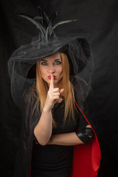 Woman dressed in a witch costume with a black feathered hat and a red lined cape, making a shush gesture with a finger on her lips