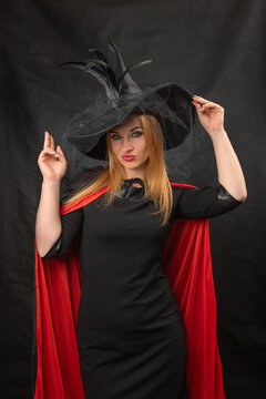 Young woman confidently posing as a witch, wearing a black dress, a red cape, and a black hat with feathers and veil, representing mystery and enchantment