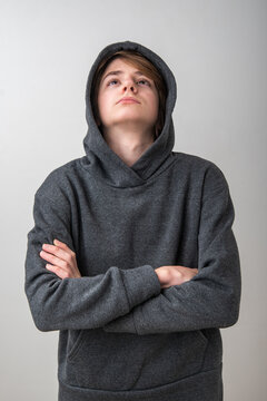 Young boy in a gray hoodie with crossed arms gazes upward in quiet contemplation, conveying dreaming, hope, concentration and future aspiration against a plain studio background