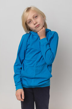 Young blonde boy standing against a plain backdrop, wearing a casual blue hoodie and dark pants, contemplating with one hand resting under his chin