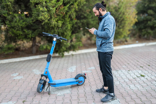 Man Stands Beside Blue Electric Scooter On Paved Park Path, Checking Smartphone Near Leafy Trees. Outdoor Scene Suggests Urban Mobility, Navigation, Leisure Travel, And Modern Connected Lifestyle