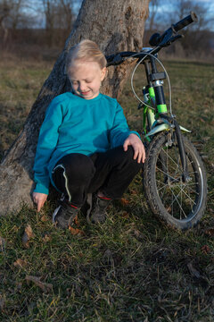 Little girl squatting by a tree on green grass with his small bicycle leaning against the trunk, exploring nature with a curious, joyful expression during outdoor play