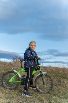 Young girl with blonde braids standing beside her neon green bicycle on a grassy hill, wearing a polka dot jacket and activewear, looking at the viewer