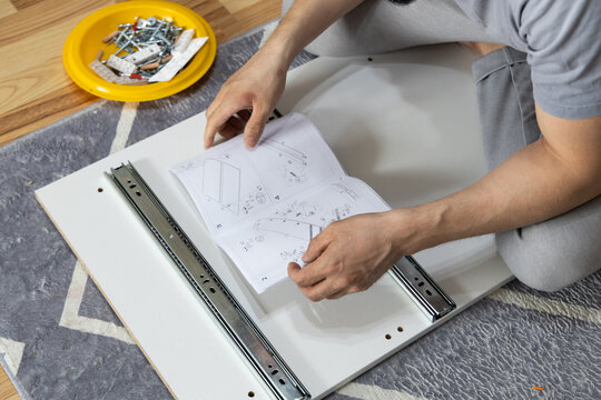 Close View Of Person Assembling Flat Pack Furniture On Patterned Rug, Reading Printed Instructions While Aligning Drawer Slides, Wood Panel, Screws And Hardware Parts During Home Improvement