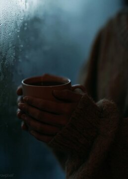 Person's hands holding a cup of coffee. the person is wearing a brown sweater and is standing in front of a window with raindrops on it. the background is blurred, but it appears to be a cloudy sky.