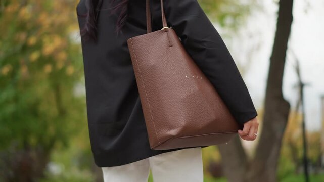 Close up of woman walking outdoors while holding and carrying brown leather tote bag. subject wears dark clothing with focus on her side profile as woman moves through urban park setting