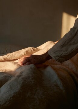 Person's hand resting on a bed. the bed is covered with a beige-colored blanket and there is a pillow visible on the right side of the image. the background is a plain white wall.