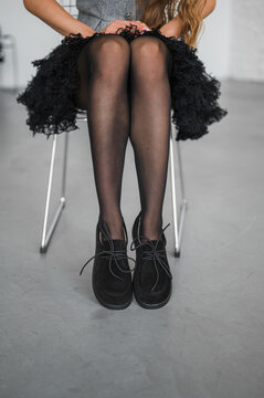 Seated woman in black sheer tights, ruffled skirt, and lace up shoes, crossed knees visible in cropped fashion portrait with neutral studio background and soft light
