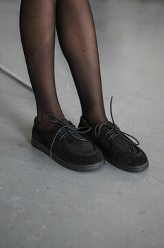 Cropped fashion portrait of seated woman showing black sheer tights, ruffled skirt, and lace up shoes, with crossed knees, resting hands, neutral studio floor, metal chair legs, and soft even light