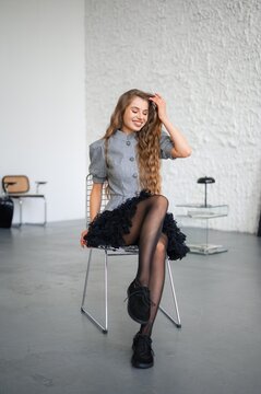 Young woman with long wavy hair smiling while seated in modern studio chair wearing grey jacket and black skirt, with woman, smile, portrait, seated detail, showing hair, longhair, wavyhair, fashion