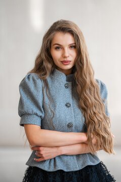Close portrait of young woman with long wavy light brown hair and natural makeup wearing grey wool coat with decorative buttons, arms crossed gently across waist, soft neutral background and