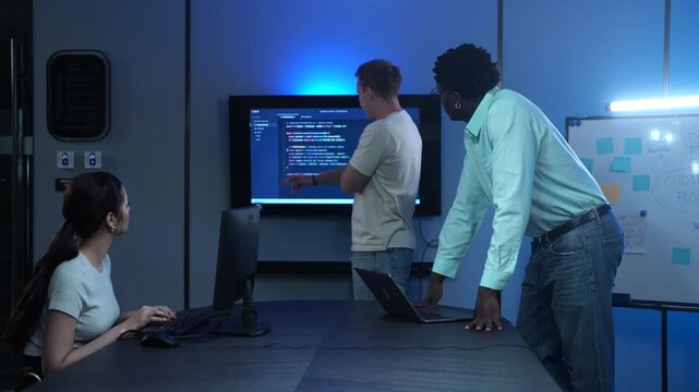 Diverse software developer team analyzing code on large monitors in dark IT office. Asian female and African American male programmers discussing technology projects. Corporate cybersecurity teamwork