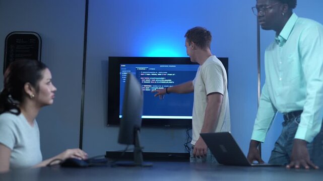 Diverse software developer team analyzing code on large monitors in dark IT office. Asian female and African American male programmers discussing technology projects. Corporate cybersecurity teamwork
