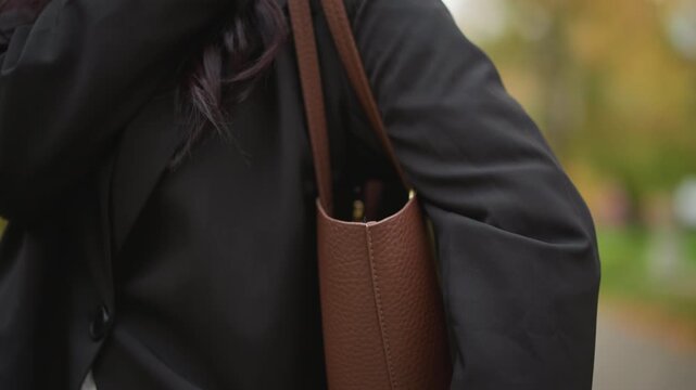 Close up of woman holding and adjusting brown leather tote bag with FASHION embossed on it. subject wears black coat white pants and carries handbag in outdoor park setting with soft natural